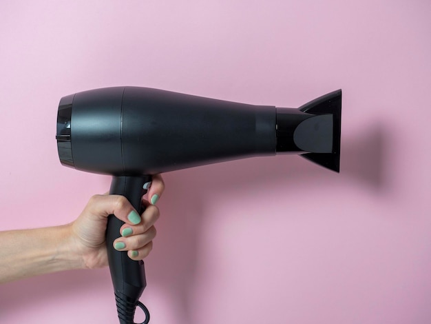 Black hair dryer in a woman's hand on a pink background. Hair Care Product.