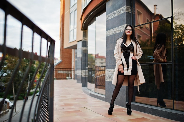 Black hair sexy woman in glasses and coat posed against building with modern windows