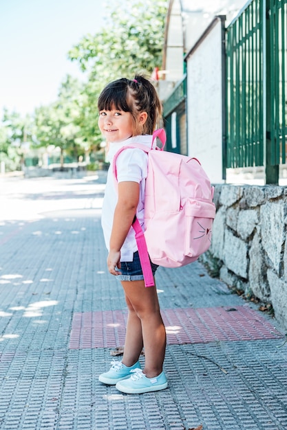 Foto bambina dai capelli neri che indossa uno zaino rosa per strada