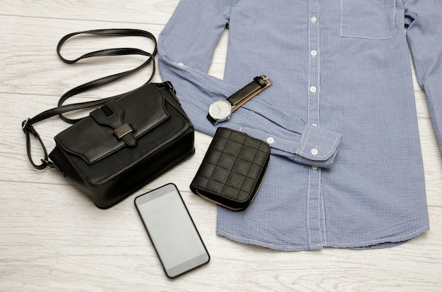 Black handbag, mobile phone, purse and a watch in the blue shirt. Fashion concept. top view