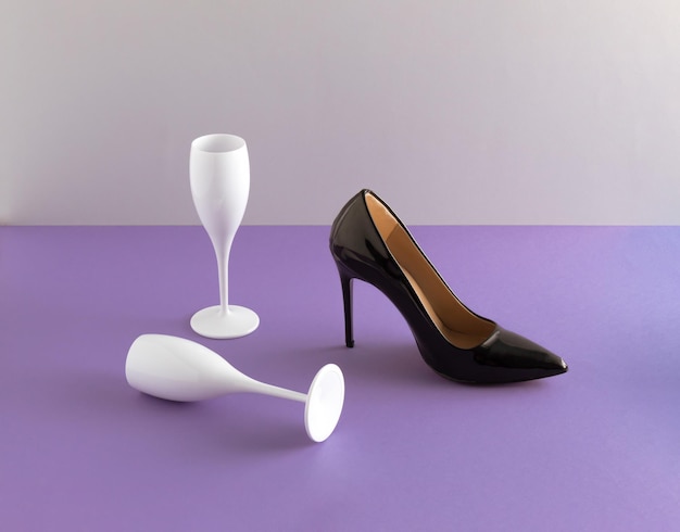 Foto scarpa nera con tacco alto e bicchieri di champagne bianchi su sfondo viola idea creativa per una festa minima