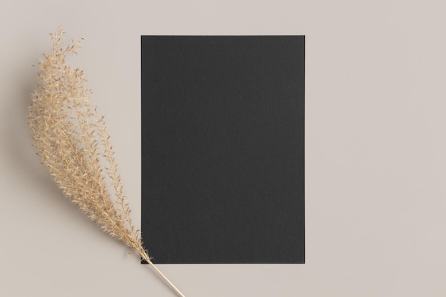 Black invitation card mockup with a dried grass decoration on a beige table 5x7 ratio similar to A6 A5