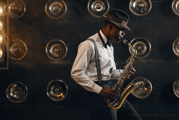 Black jazzman in hat plays the saxophone on the stage with spotlights. Black jazz musician preforming on the scene