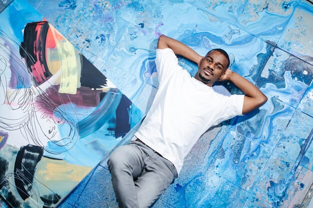 Black male artist lying on blue paint stained floor next to an abstract painting