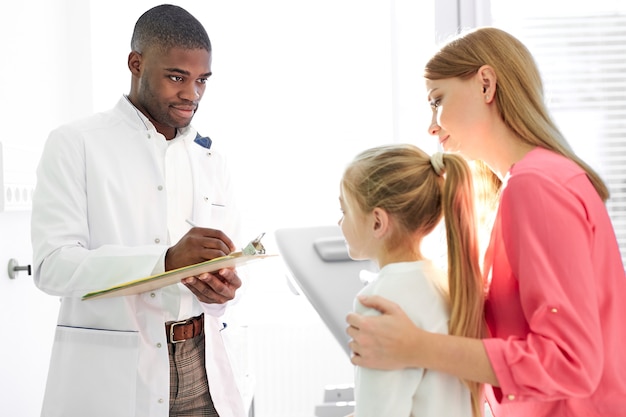 Black male doctor talking to child and his mother during health checkup at the clinic, mom and girl get consultation by professional pediatrician or general practitioner during visit to the hospital