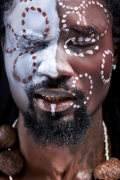 Black male feeling pain, stand with closed eyes isolated in studio, having paintings drawings on face, ethnic national make-up