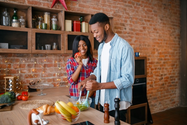 Black man cooking on kitchen, wife drinks coffee