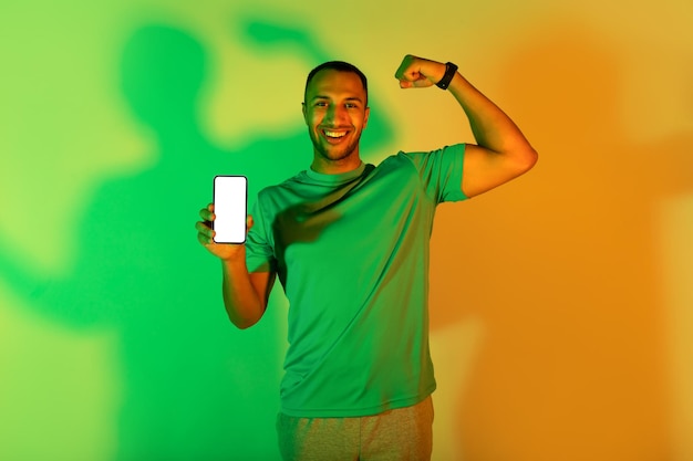 Black man demonstrating smartphone screen and biceps muscles in studio