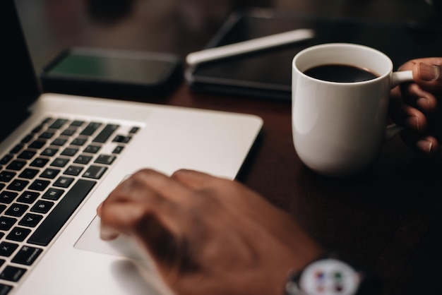 Black man drinking coffee at working at a computer desk with smartphone and laptop