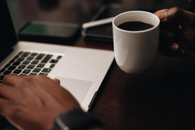 black man drinking coffee at working at a computer desk with smartphone and laptop