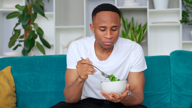 Black man eating healthy salad sitting indoors on the sofa Healthy eating concept