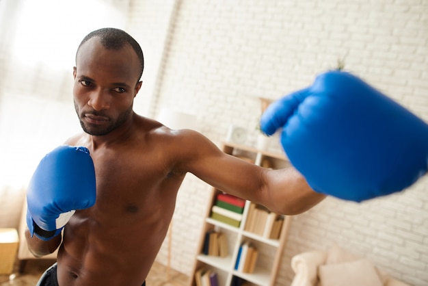 Black man is boxing in sporty gloves at home.