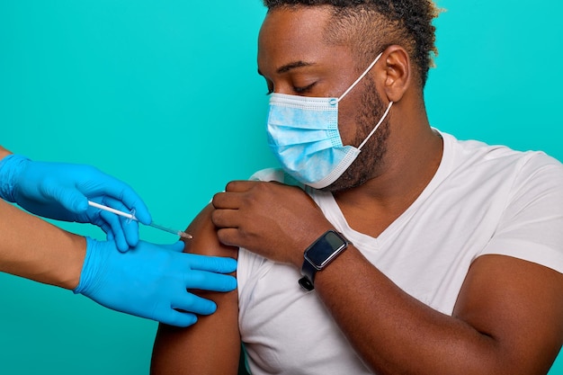 L'uomo di colore in una maschera guarda attentamente come le mani di un medico in guanti blu vengono iniettate con una siringa con un vaccino contro il coronavirus per l'immunizzazione contro covid19