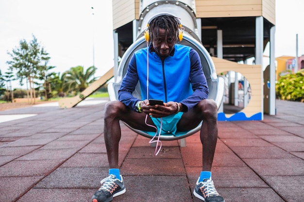 black man sitting in a playground in sportswear,listening to music with headphones through his phone