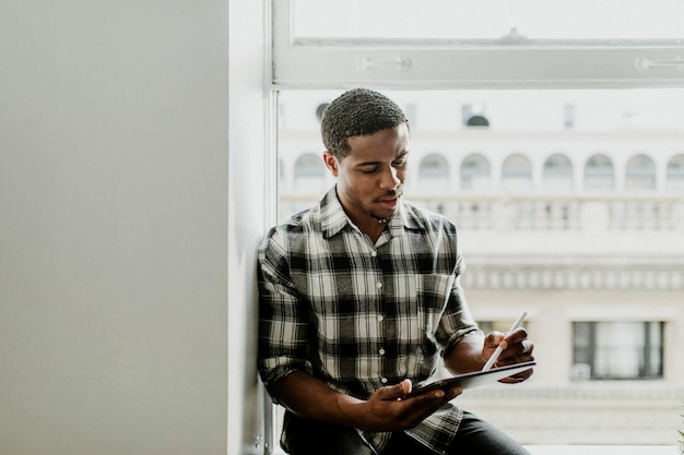 Black man using a stylus to draw on his digital tablet by the window