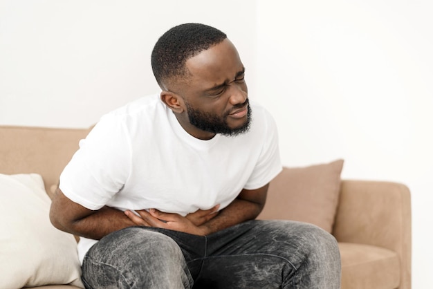 Black man with abdominal pain suffering from painful abdominal cramp in stomach sitting on couch at home Abdominal pain stomach inflammation and appendicitis concept