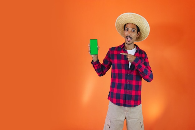 Black Man With Junina Party Outfit holding a tablet or mobile Isolated on Orange Background Young man wearing traditional clothes for Festa Junina Brazilian June festival