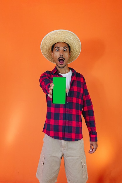 Black Man With Junina Party Outfit holding a tablet or mobile Isolated on Orange Background Young man wearing traditional clothes for Festa Junina Brazilian June festival