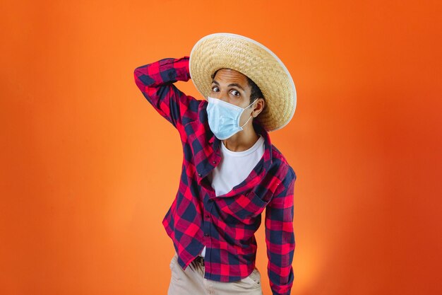 Black Man With Junina Party Outfit and pandemic mask Isolated on Orange Background Young man wearing traditional clothes for Festa Junina Brazilian June festival