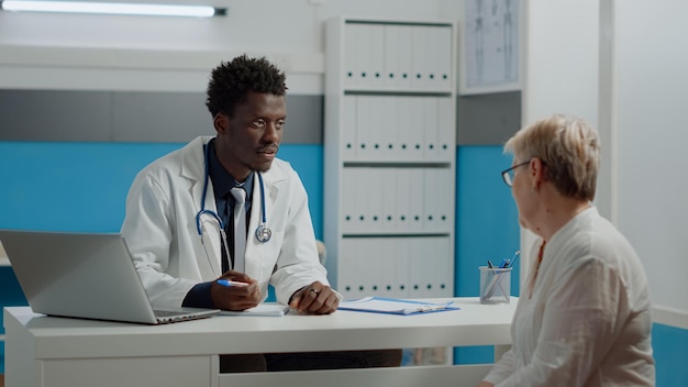 Black man working as doctor talking to senior woman in healthcare office. Medic of african american ethnicity consulting old patient about medical treatment and medicine at modern facility