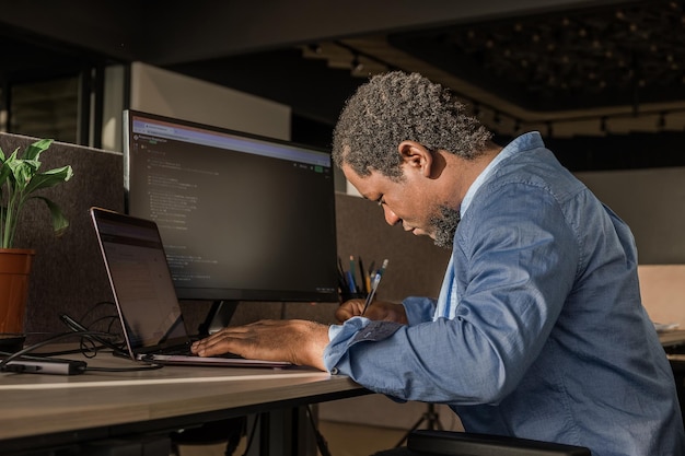 Black man writing lines of code on desktop pc with two monitors and a laptop aside in stylish office