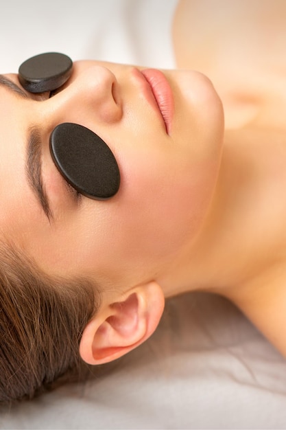 Black massage stones lying on the eyes of the young caucasian woman. Facial massage in a spa.