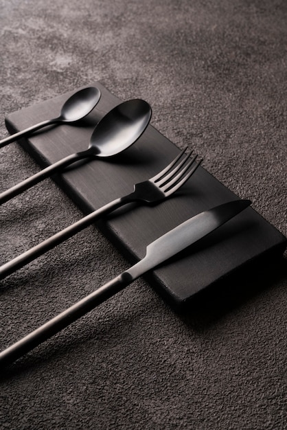 Black matte cutlery set-fork, spoon, knife, on dark concrete. Minimalistic still life, stylish tableware. vertical photo close-up.