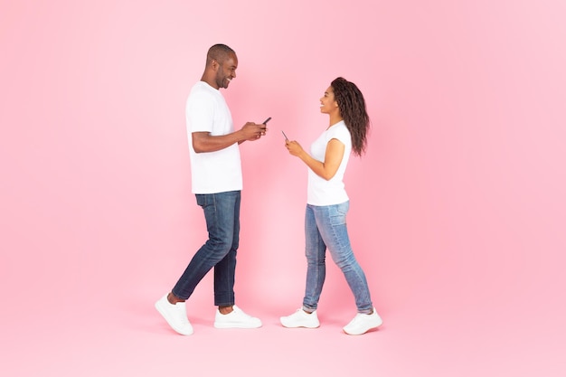 Black middle aged man and young woman using smartphones standing over pink studio background full length side view