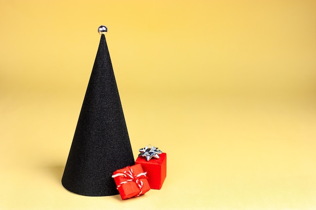 写真 黄色の背景のポップアートに赤い贈り物と円錐形の黒いミニマルなクリスマスツリー...