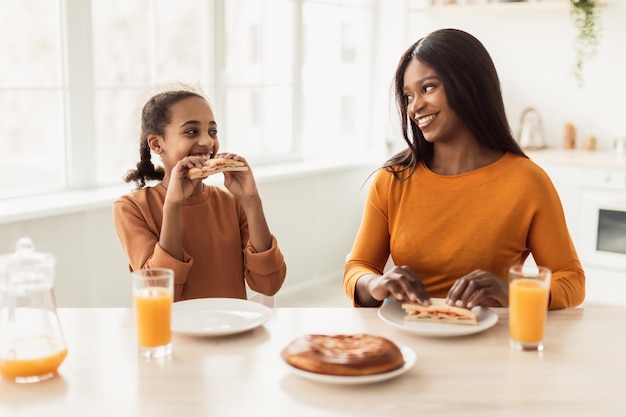 Black mom and daughter eating sandwiches having lunch in kitchen