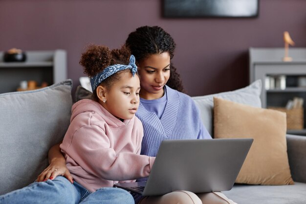 Photo black mother and daughter using laptop sitting on sofa together