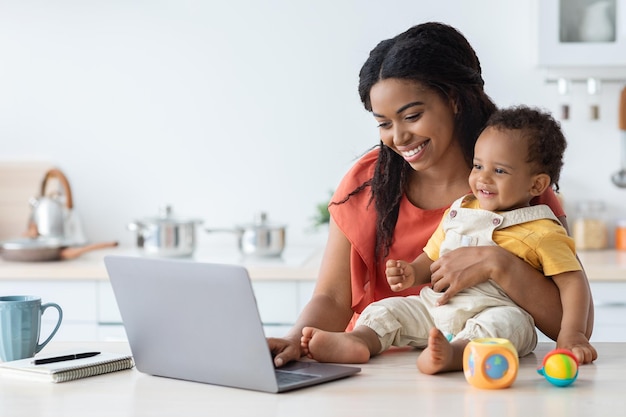 Madre nera che si prende cura del bambino e lavora al computer portatile a casa