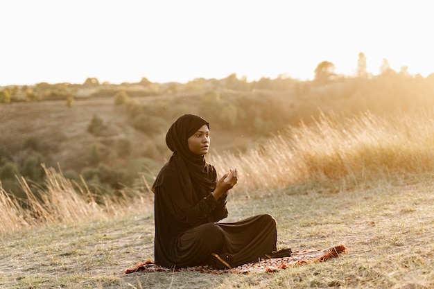 Photo black muslim woman praying to allah at sunset. solat traditional praying to god on the carpet.