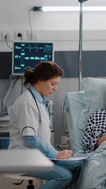 Black nurse checking patient vitals monitoring heart rate injecting vitamin in iv fluids drip bag in hospital ward