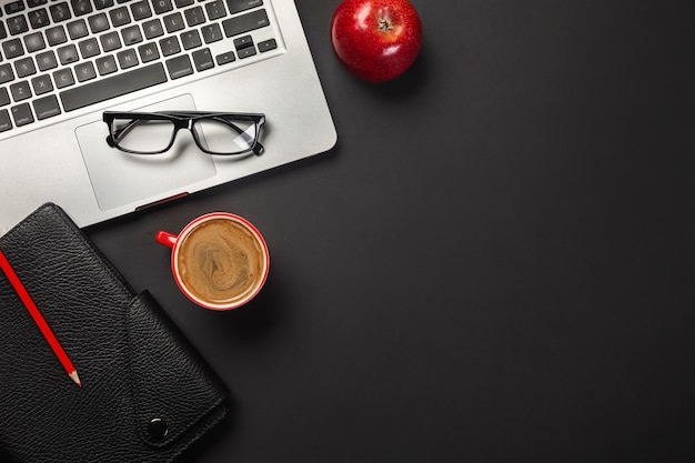 Black office desk table with blank screen laptop computer, notebook, cup of coffee, red apple and other office.Top view with copy space.