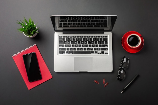 Black office desk table with blank screen laptop computer, notebook, mobile phone and red cup of coffee. Top view.