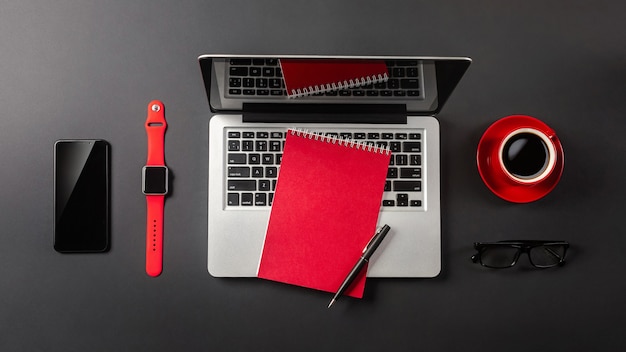 Black office desk table with blank screen laptop computer, notebook, watch, mobile phone and red cup of coffee. Top view.