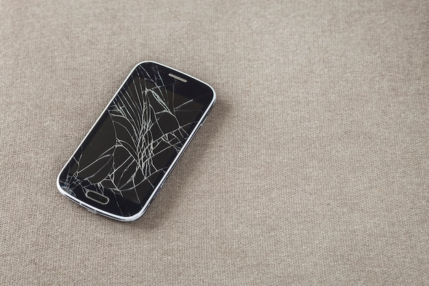 Black old cellphone with cracked screen on light cloth copy space background. Gadget repair and maintenance concept.