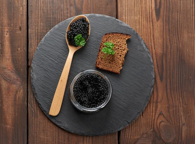 Photo black paddlefish caviar in a wooden spoon a slice of bread and a jar on a black round graphite