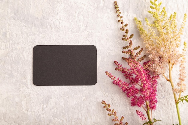 Black paper business card mockup with purple astilbe flowers on\
gray concrete background top view copy space