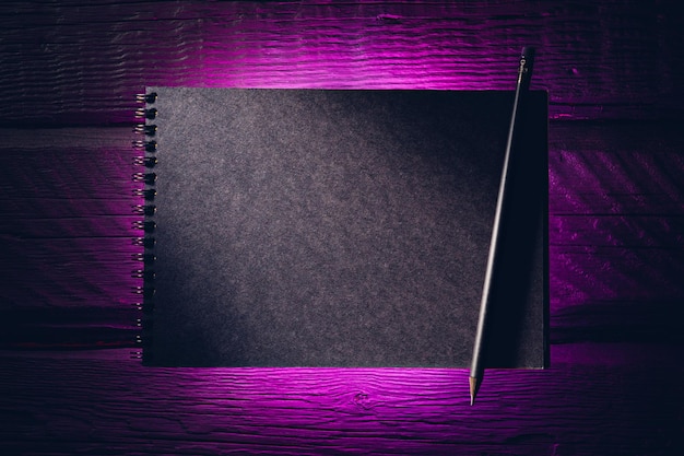 Black paper notebook on black wooden planks in purple neon light