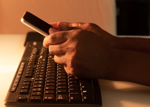 Черный человек держит мобильный телефон на клавиатуре на белом столе с теплым светом