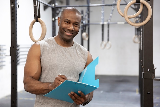 Black personal trainer in fitness center. He is smiling and taking notes in a notebook. Space for text.