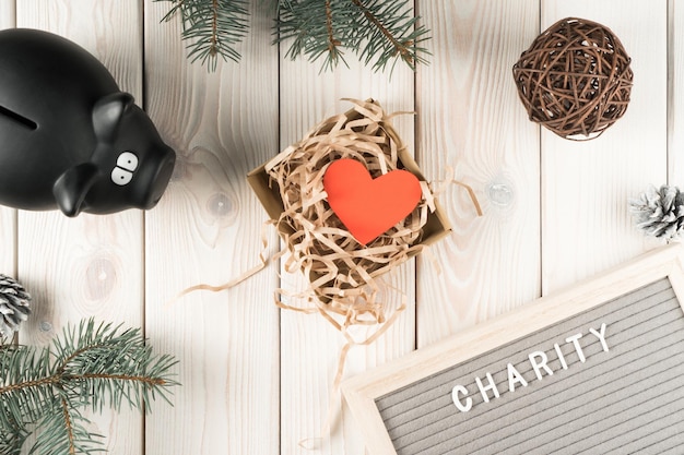 Black piggy bank for donation and present box with red paper heart postcard top view Decorative spruce branches on white planking Giving money Kindness and Christmas or New Year charity concept