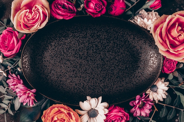 Black plate frame decorated with beautiful summer flowers. Pink roses and white daisies top view, toned photo.