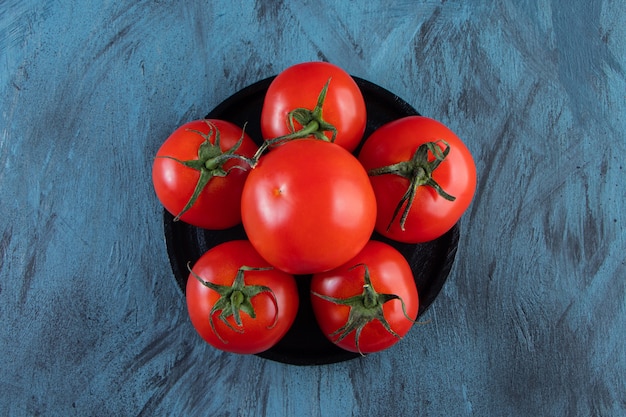 Black plate of red fresh tomatoes on blue surface.