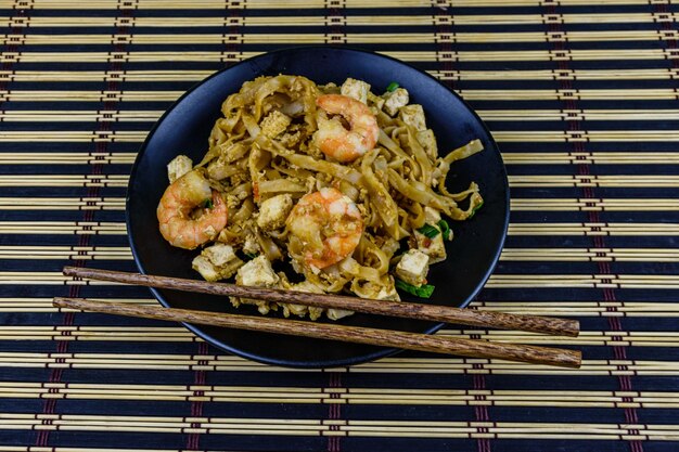 Black plate with pad thai and chopsticks on a bamboo mat Top view