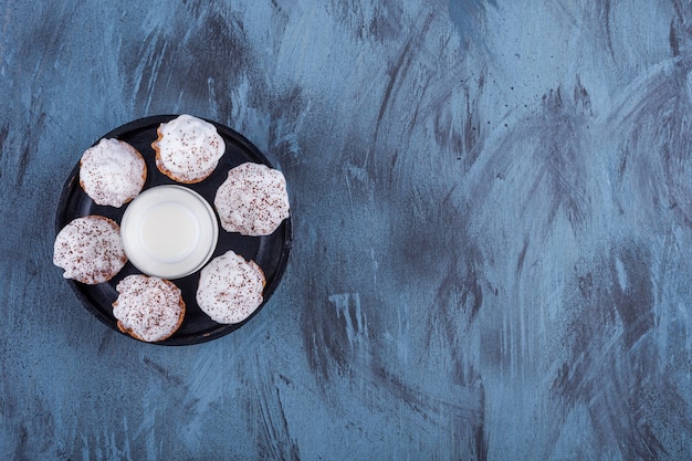 Black plate with sweet creamy cupcakes and glass of milk on marble.