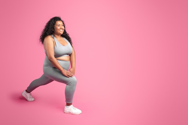 Black plus size woman training stretching legs and making lunges pink background free space full