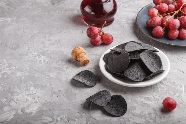 Black potato chips with charcoal, balsamic vinegar in glass, red grapes on a blue ceramic plate on a gray concrete surface. side view.